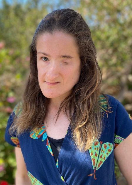 A woman with long brown hair stands outdoors in front of a blurred background of greenery and pink flowers. She is wearing a navy blue jumpsuit with a colourful print and looks calmly toward the camera.