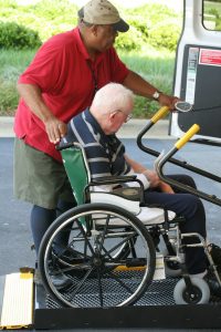 A taxi driver helps an older gentleman using a wheelchair to board a taxi.