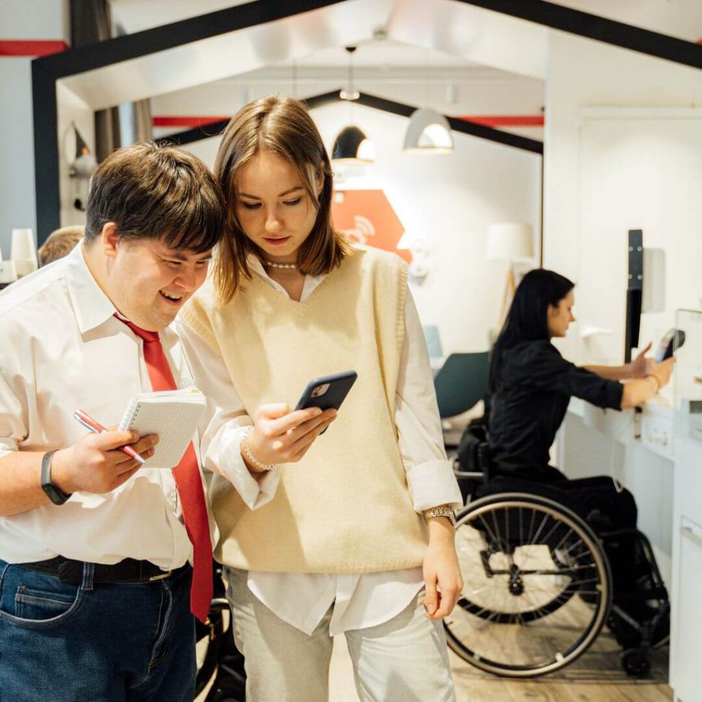 Two people stand looking at a mobile phone. A person in a wheelchair is visible in the background holding a device.