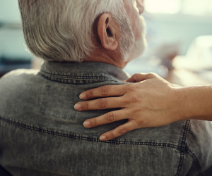 Older man picture from behind with his face slightly turned to the side. There is a hand resting on his shoulder. He has white hair, a beard and is wearing a grey shirt.