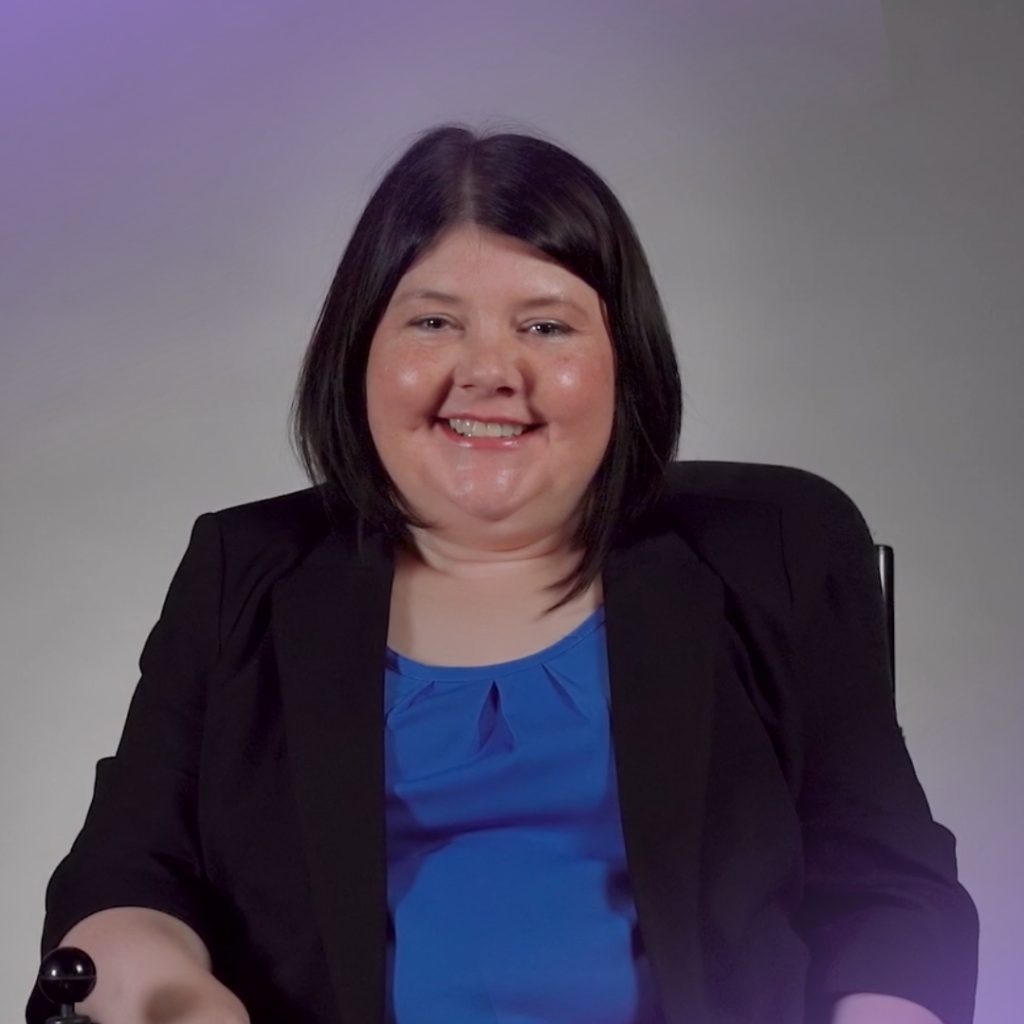 A woman with straight dark hair smiles at the camera while seated in a wheelchair. She is wearing a royal blue blouse under a black blazer, and the background is a neutral grey with soft purple lighting effects in the corners.