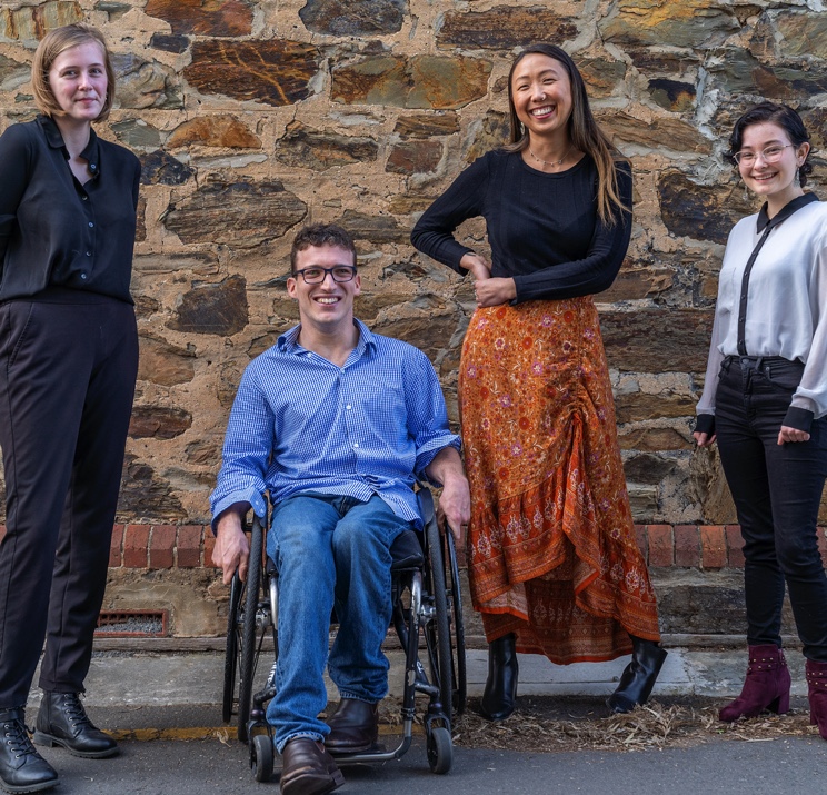 Four young people in front of a brick wall. Three females are standing and one male is in a wheelchair.