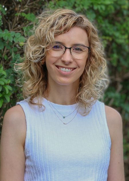 A smiling woman with shoulder length, curly, blonde hair is wearing glasses and a sleeveless white top. There is greenery in the background.