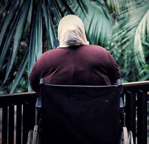 Photo of older woman in a wheelchair photographed from behind. It looks like she is on a balcony looking out at some palm trees. She is wearing a deep red cardigan.