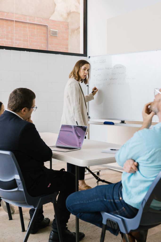 A person is writing on a white board and there are people sitting at a table facing the whiteboard.