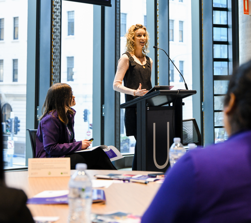 A presenter stands wearing a long sleeved top and a black vest.