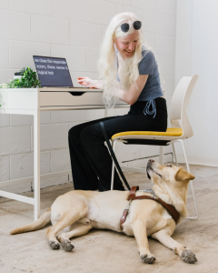 Alana sits at a desk with her golden guide dog, Rocket, laying at her feet. They're both looking at each other.