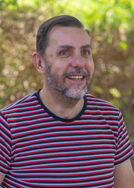 A man with short dark hair and a salt-and-pepper beard smiles. He wears a red, black, and white striped t-shirt. Behind him, the background is softly blurred with sunlit trees and greenery.