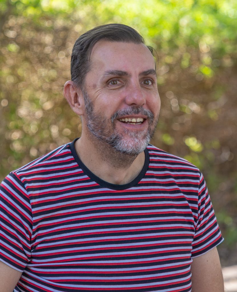 A man with short dark hair and a salt-and-pepper beard smiles. He wears a red, black, and white striped t-shirt. Behind him, the background is softly blurred with sunlit trees and greenery.