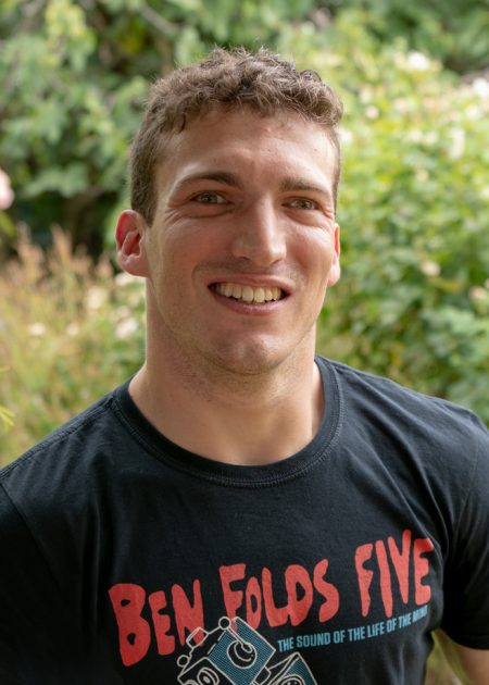 A man with short, wavy brown hair smiles to camera. He is wearing a black "Ben Folds Five" t-shirt with red lettering. The background includes blurred plants and flowers.