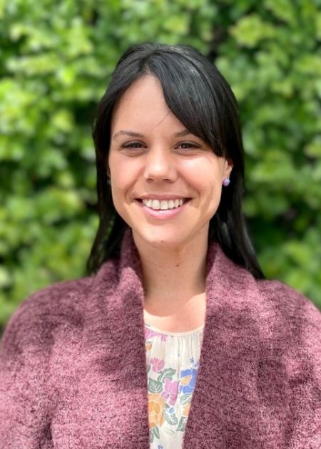 Ayla, a woman with long dark hair, is standing in front of blurred greenery and smiling at the camera. She is wearing a brown cardigan over a floral top.