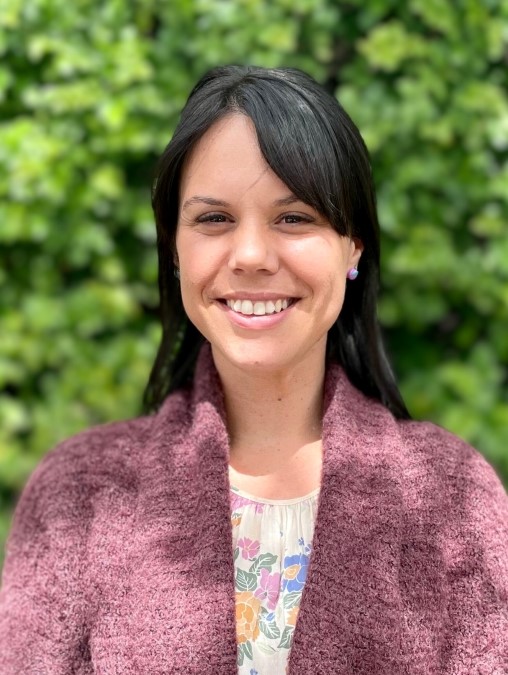 Ayla, a woman with long dark hair, is standing in front of blurred greenery and smiling at the camera. She is wearing a brown cardigan over a floral top.