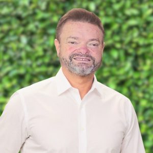 A man with short hair and a trimmed beard smiles, wearing a white button-up shirt against a leafy green background.