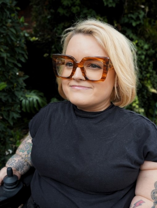 A woman with chin-length blonde hair smiles gently while looking off to the side. She is wearing oversized tortoiseshell glasses and a black t-shirt, with tattoos visible on both arms. Her hand rests on the control of a power wheelchair. There is blurred greenery behind her.