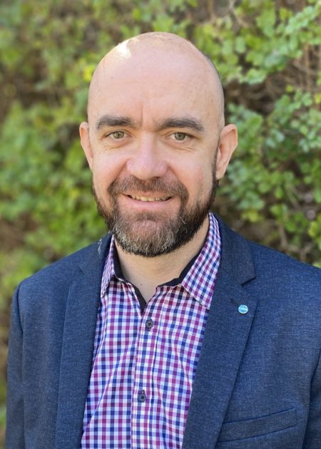 Photo of Brad, who is smiling at camera. He is bald and has a beard, and is wearing a navy blazer with a checked shirt. There is blurred greenery in the background.