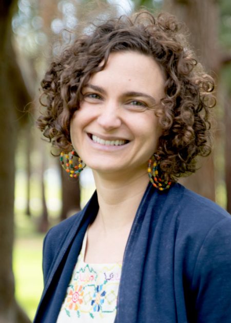 A woman with short, curly hair smiles while standing outdoors. She is wearing large, colorful beaded earrings, a navy blue cardigan, and a light top with a bright, embroidered floral pattern. The background features blurred trees and green grass.