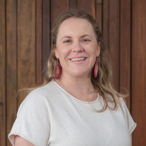 Danelle smiles at the camera. She has long light-brown hair and is wearing a light top and colourful earrings, against a wooden background.