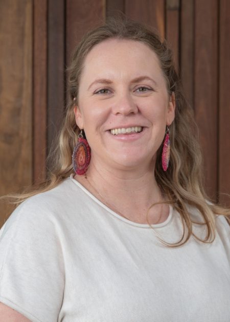 Danelle smiles at the camera. She has long light-brown hair and is wearing a light top and colourful earrings, against a wooden background.