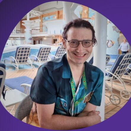 A smiling Daniel is seated at a table with sun loungers in the background. He is wearing a patterned, blue short sleeve shirt. He has short dark hair and wears black glasses.