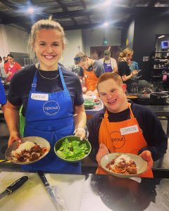 2 people stand, smiling, proudly showing off food created at a peer network. A busy kitchen behind them with many people cooking.