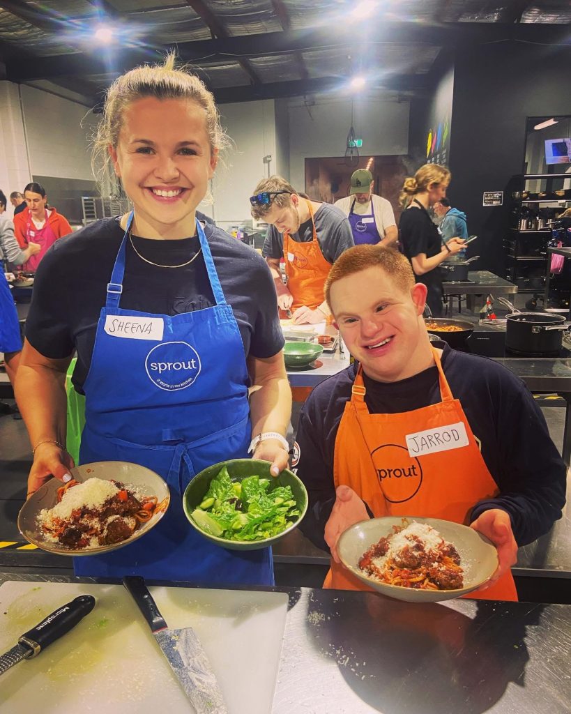 2 people stand, smiling, proudly showing off food created at a peer network. A busy kitchen behind them with many people cooking.