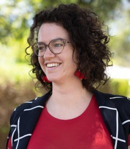 A person with short, curly dark hair smiles while standing outdoors in bright natural light. They are wearing glasses, red cherry-shaped earrings, a red top, and a black jacket with a white grid pattern. The background is blurred greenery.