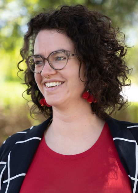 A person with short, curly dark hair smiles while standing outdoors in bright natural light. They are wearing glasses, red cherry-shaped earrings, a red top, and a black jacket with a white grid pattern. The background is blurred greenery.