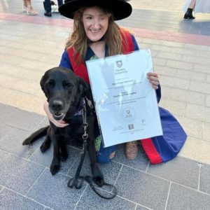 Ellen simles, crouching next to Inka with an arm around her. Ellen wears a doctoral robe and holds her graduation certificate.
