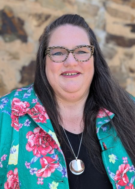 Heidi, a woman with long dark hair wearing dark framed glasses, is smiling at the camera. She is standing in front of a stone wall and is wearing a green floral jacket over a black top.