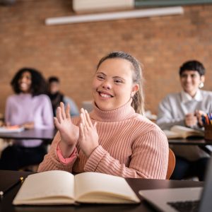 Girl sitting behind a desk with an open book in front of her. She is smiling and has her hands up in front of her as though she is about to clap them. She is looking forward and smiling.