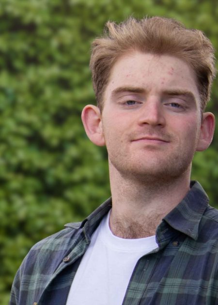Young man with short reddish hair in a green plaid shirt and white tee, standing before a leafy green background.