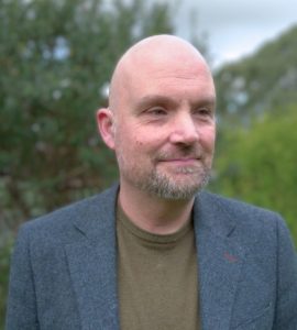 A man with a bald head and short beard smiles slightly while looking off to the side. He is wearing a bluish grey blazer over an olive green top. The background is outdoors with blurred greenery and a cloudy sky.