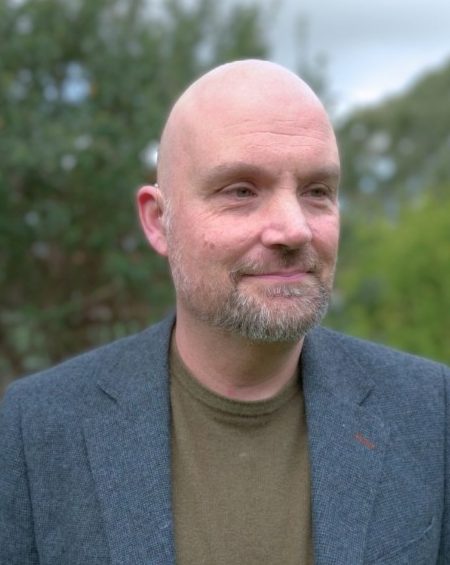 A man with a bald head and short beard smiles slightly while looking off to the side. He is wearing a bluish grey blazer over an olive green top. The background is outdoors with blurred greenery and a cloudy sky.