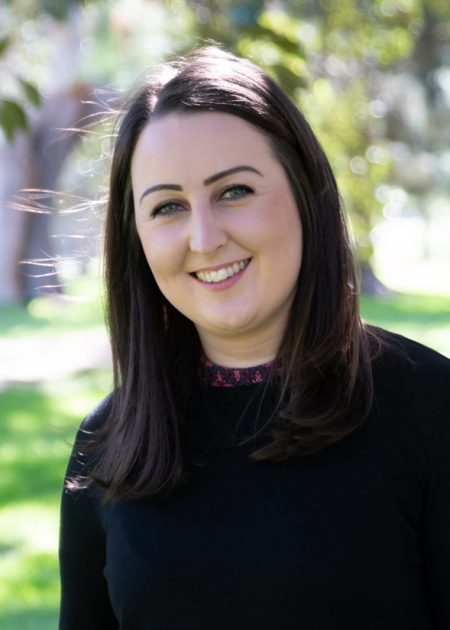 A woman with straight, dark brown hair smiles while standing outdoors. She is wearing a black long-sleeved top. The background is filled with green grass, trees, and dappled sunlight.
