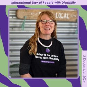 Dark purple and light green background with a square frame containing photo of Regional Connector, Liluella. She is wearing a black Purple Orange t-shirt, glasses and a white shell necklace. She has long red hair. Small text around border reads: International Day of People with Disability. 3 December 2024.