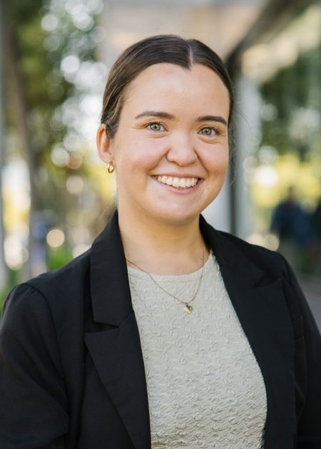 A young woman with dark hair tied back smiles while standing outdoors in a city setting. She is wearing a light textured top, a black blazer, and a gold necklace. Trees and a glass building line the footpath in the background.