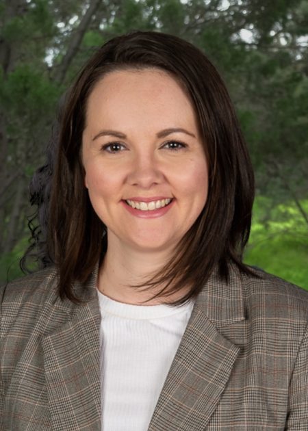 A woman with straight, dark brown hair smiles at the camera while standing outdoors. She is wearing a grey plaid blazer over a white top. The background features softly focused green trees and foliage.