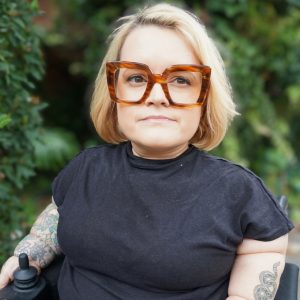 Belle Owen pictured outside with blurred greenery in background. Belle has short blonde hair and wears large brown framed glasses.