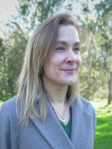 A woman with straight, blonde hair smiles gently while looking off to the side. She is wearing a grey coat over a green top, with a heart-shaped pendant necklace and green earrings. The background is a sunlit park with trees and grass in soft focus.