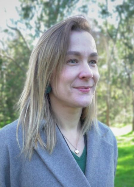 A woman with straight, blonde hair smiles gently while looking off to the side. She is wearing a grey coat over a green top, with a heart-shaped pendant necklace and green earrings. The background is a sunlit park with trees and grass in soft focus.