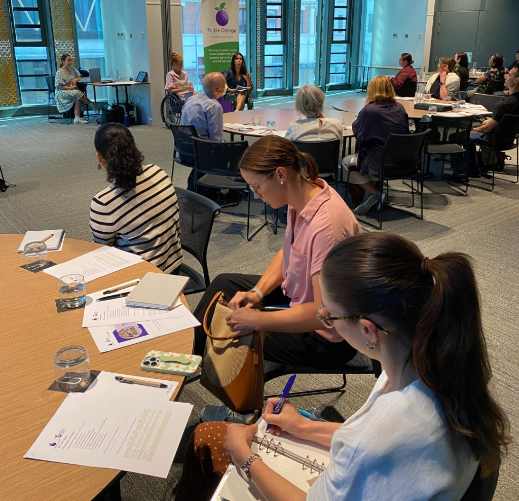 A group of people sit, listening to presenters and writing notes.