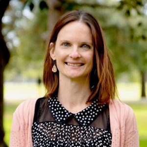 A woman with straight, shoulder-length brown hair smiles at the camera. She is wearing drop earrings, a black top with white polka dots, and a light pink cardigan. The background is blurred greenery with trees and sunlight.