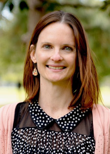 A woman with straight, shoulder-length brown hair smiles at the camera. She is wearing drop earrings, a black top with white polka dots, and a light pink cardigan. The background is blurred greenery with trees and sunlight.