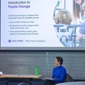 A woman in a bright blue top sits and smiles during a presentation in a modern conference room. Behind her, a large screen displays a slide titled "Introduction to Purple Orange" with key points about supporting people with disability. The slide includes a photo of people interacting, including a wheelchair user, and a play button labeled “Play Video (Why Purple Orange?).”