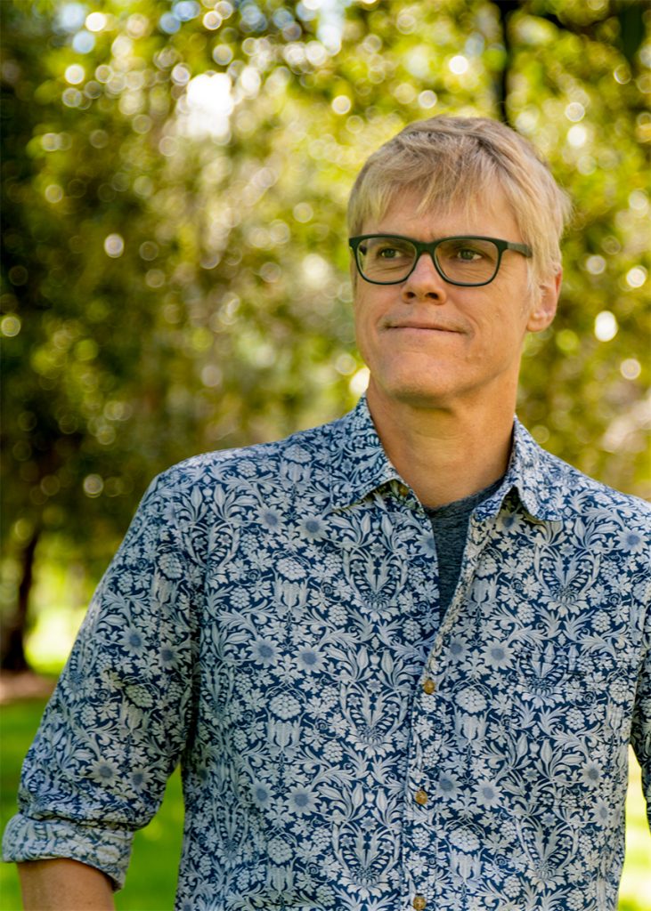 A man with short blond hair and black-rimmed glasses stands outdoors, looking slightly to the side with a calm expression. He is wearing a blue and white patterned shirt. The background is filled with sunlit trees and blurred greenery.