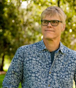 A man with short blond hair and black-rimmed glasses stands outdoors, looking slightly to the side with a calm expression. He is wearing a blue and white patterned shirt. The background is filled with sunlit trees and blurred greenery.