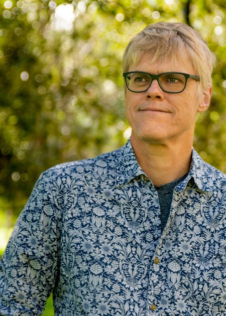 A man with short blond hair and black-rimmed glasses stands outdoors, looking slightly to the side with a calm expression. He is wearing a blue and white patterned shirt. The background is filled with sunlit trees and blurred greenery.