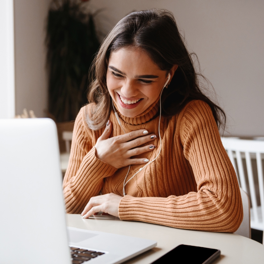 A person sits at a table looking at a laptop which is slightly blurred in the foreground. They are wearing an orange long sleeved turtleneck and have long dark hair.