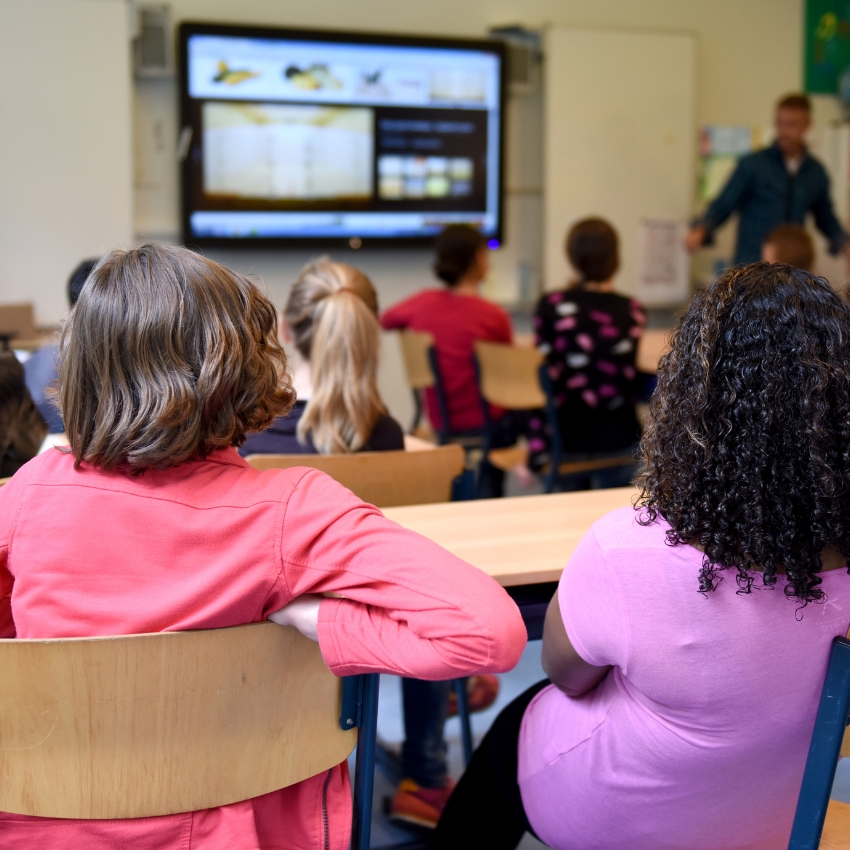 Students sitting in a classroom with the teacher visible at the front. There is a blurred large screen in front of the class and students can be seen at their desks from behind.