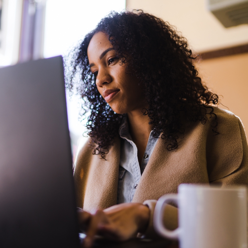 A woman with long dark curly hair sits at a desk looking with interest at a computer monitor directly in front of her.